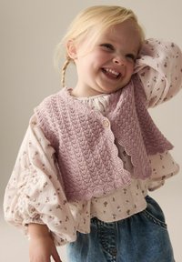 Lichtroze gebreide vest met een gegolfde rand, opgedragen over een bloemenblouse. Losse mouwen en denim onderkleding maken de outfit compleet.