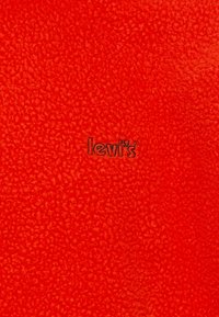 Červená texturovaná látka s jemným vzorem a černým logem "Levi's" v pravém dolním rohu.