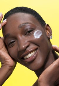 Modelka z krótkimi, teksturowanymi czarnymi włosami, uśmiechająca się. Ma na policzku nałożoną białą kremową substancję, a w tle znajduje się żółta ściana. Naturalny odcień skóry i zdrowe zęby.