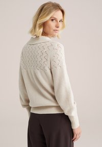 Svetlo bež pulover z rebrasto teksturo in čipkastim hrbtom. Ima ovratnik in sproščen kroj, v kombinaciji s temno rjavimi hlačami.