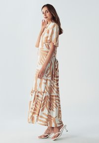 Dlhé, voľné šaty v krémovej farbe s hnedým abstraktným vzorom, krátkymi rukávmi, vrstveným spodným okrajom a voľným strihom, skombinované s bielymi sandálmi na vysokom opätku.