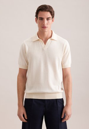 Homme portant une chemise en maille crème à manches courtes avec col et un pantalon foncé, debout devant un fond neutre clair.