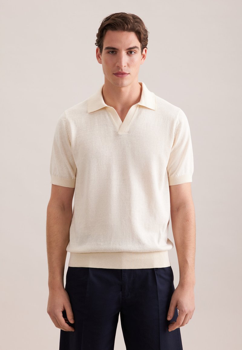 Homme portant une chemise en maille crème à manches courtes avec col et un pantalon foncé, debout devant un fond neutre clair.