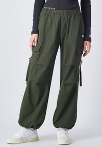 Olijfgroene cargobroek met een elastische taille. Beschikt over oversized opgestikte zakken, trekkoordaccenten en taps toegesneden uiteinden. Gecombineerd met witte sneakers.