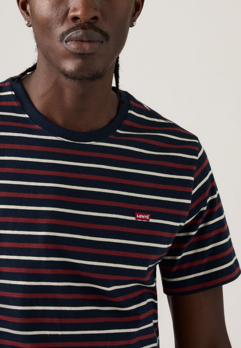 Mann trägt ein Levi's T-Shirt mit kurzen Ärmeln in Marineblau, Weiß und Burgunderrot gestreift, mit kleinem rotem Levi's Logo auf der Brust.