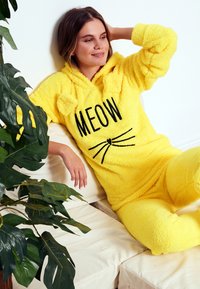 Salopetă din fleece galben cu glugă, având textul "MEOW" și grafica unor mustăți. Textură moale și croială lejeră.