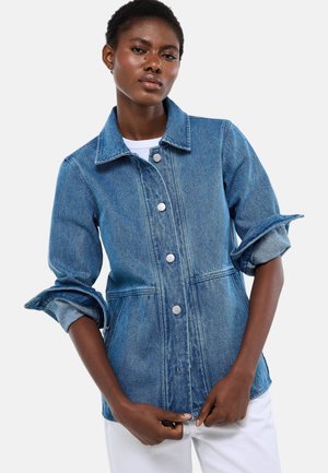 Femme portant une veste en jean bleue avec les manches retroussées et des boutons blancs, associée à un pantalon blanc, sur un fond clair uni.