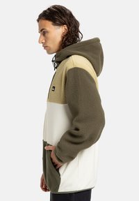 Fleece mikina s kapucí v olivově zelené, krémové a béžové barvě. Obsahuje kapuci, přední kapsu a nastavitelnou šňůrku. Měkká textura s oversized střihem.