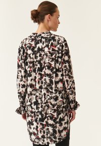 Blusa de manga larga en blanco con un patrón abstracto en negro y rojo. Presenta un corte holgado, puños con botones y un detalle de yugo en la parte posterior.