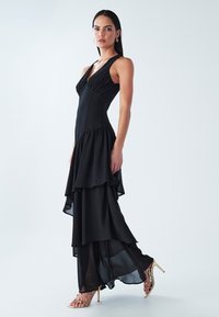 Juoda sluoksniuota suknelė su plona silueto linija, giliu V formos iškirpte ir laisvai krentančia sijono dalimi. Papildyta auksinėmis basutėmis su dirželiais. Lygus audinys.