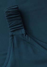 Turkusowy elastyczny materiał z marszczoną linią dekoltu i gładką teksturą. Posiada płaskie szwy i dopasowany krój, co zwiększa komfort i elastyczność.