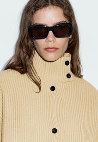 Beige ribbad tröja med hög krage och svarta knappar. Grov textur; modell som bär mörka rektangulära solglasögon.