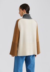 Sweater coloré avec des sections grises, crème, marron et bleues. Il présente un col monté et une texture côtelée sur les manches et le bas.