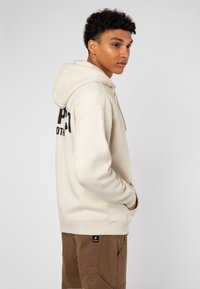 Cream hoodie with a front pocket and large black graphic text on the back. Soft texture, with a drawstring hood and relaxed fit. Brown cargo pants.