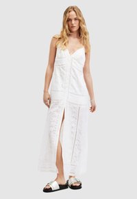 Weißes Maxikleid mit tiefem V-Ausschnitt, bestickten Spitzenverzierungen und einem seitlichen Schlitz. Das Model trägt schwarze Rutschsandalen mit silbernen Akzenten.