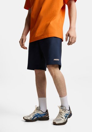 Mężczyzna w pomarańczowej koszulce, granatowych spodenkach z logo "PROSTO", białych skarpetkach i beżowych butach do biegania z niebieskimi akcentami, stojący na białym tle.