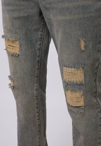 Jean en denim bleu délavé avec plusieurs patchs abîmés aux bords effilochés et des accents tissés. Coupe large avec un design décontracté.