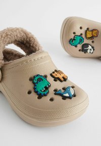 Sabots beiges avec un intérieur douillet. Dotés de charms sur le thème des animaux : dinosaure, tigre, baleine et panda. Des trous de ventilation sur le dessus.