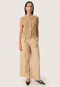 Beige stickad ärmlös topp med knappstängning, kombinerad med beiga vida byxor; modellen bär svarta spetsiga ballerina-skor.