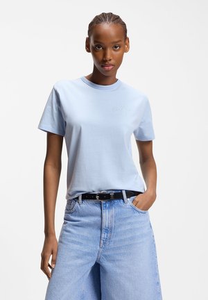 Vrouw met gevlochten haar, een lichtblauw T-shirt dat in een lichtblauwe, hooggesneden jeans met een zwarte riem is gestopt, staand met één hand in de zak.
