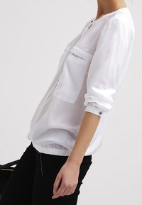 Vit långärmad blus med rund halsringning, två bröstfickor, elastisk nederkant och knapppyssel på ärmarna. Tillverkad av lätt, mjukt tyg.