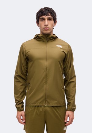 Νέος άνδρας με ελαφρύ πράσινο μπουφάν North Face με κουκούλα και αντίστοιχα σορτς, στέκεται μπροστά σε απλό φωτεινό φόντο.