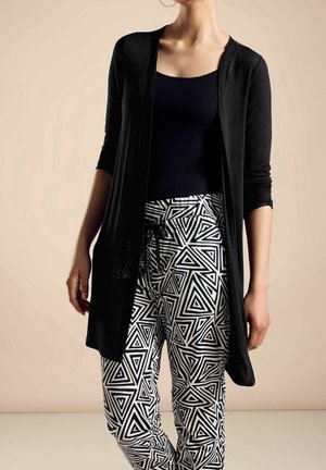 Femme portant un cardigan noir ouvert à manches trois-quarts, un débardeur noir et un pantalon blanc avec un motif géométrique triangulaire noir audacieux.
