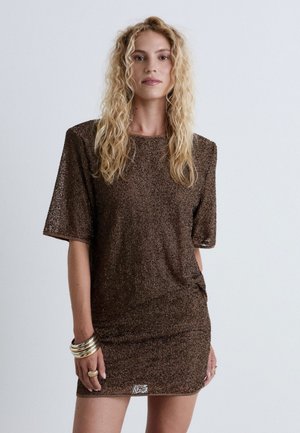 Femme aux longs cheveux blonds bouclés, portant une mini-robe marron texturée à manches courtes et des bracelets dorés au poignet gauche.