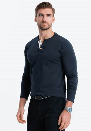 Marineblauw langslavige henleyshirt met een knoopsluiting voorzien van een witte accenten en houten knopen; gecombineerd met zwart geblokte broek.