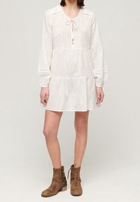 Kvinde iført en hvid langærmet kjole med teksturerede lodrette striber og brune ankelstøvler, der står foran en ensfarvet lys baggrund.