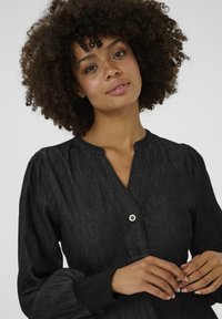 Zwarte denim overhemd met een met knopen gesloten V-hals, pofmouwen en een gestructureerde stof. Voorzien van een enkele knoopaccent bij de halslijn.