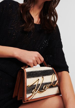 Kobieta o ciemnych włosach, ubrana w czarny, teksturowany sweter, trzyma małą paskowaną torebkę z złotym napisem "Karl" i brązową lamówką.