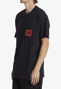 Svart bomullströja med en röd ficka på vänster sida, prydd med vit text som beskriver varumärket och designinslag.