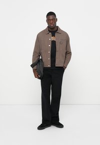 Bruine gestructureerde jacket met een knoopsluiting, gedragen over een zwart T-shirt met vlamgraphics. Zwarte broek en sneakers maken de outfit compleet.