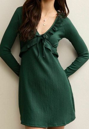 Femme portant une robe côtelée vert foncé à manches longues avec des volants et un détail noué à l'avant, debout devant un fond uni clair.