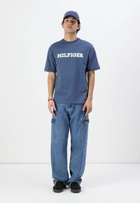 Nuori mies, jolla on päällä sininen Hilfiger-t-paita, löysät cargo-farkut, mustat lenkkarit ja siihen sopiva sininen lippalakki, seisoo tasaisen valkoista taustaa vasten.