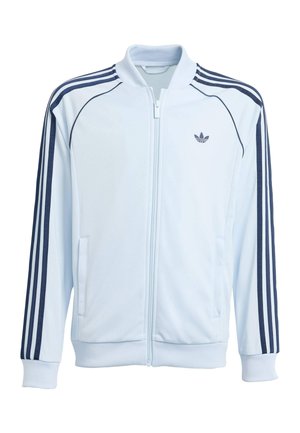 Fehér cipzáras sportdzseki tengerészkék csíkokkal az ujjain és kicsi Adidas logóval a bal mellrészen, bordázott mandzsettákkal és gallérral.