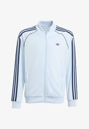 Balta sporta jaciņa ar rāvējslēdzēju, tumši zilas svītras piedurknēs un neliels Adidas logotips kreisajā krūtīs, ar ribotām aprocēm un apkakli.