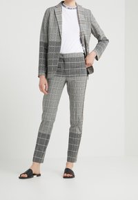 Costume à carreaux gris avec une veste ajustée et un pantalon slim. T-shirt blanc en dessous. Sandales noires à enfiler aux pieds.