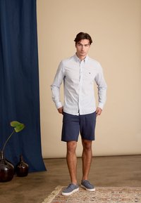 Lichtblauw overhemd met kleine witte stippen, gecombineerd met marineblauwe shorts en grijze sneakers. Staande tegen een beige achtergrond.