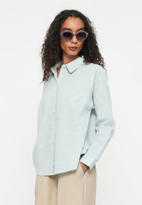 Light blue, long-sleeve shirt with a button-up front and subtle textured dot pattern. Worn with oversized, retro-style sunglasses.
