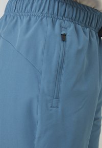 Un pantalon léger bleu avec une taille élastique et une poche latérale zippée, fabriqué dans un tissu lisse à finition légèrement texturée.