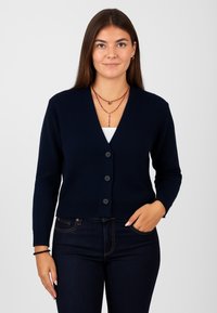 Cardigan lavorato a maglia blu navy con scollo a V, orlo a costine e cinque bottoni neri. Abbinato a una maglietta bianca e jeans scuri. Texture liscia, design aderente.