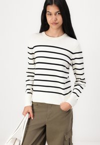 Pull blanc à rayures noires horizontales, col rond, manches longues, associé à un pantalon cargo vert olive et un sac à main blanc.
