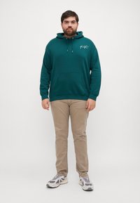 Felpa di cotone color teal con tasca frontale e cappuccio regolabile, abbinata a pantaloni beige e sneakers grigie con dettagli neri.