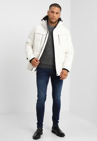 Witte pufferjack met capuchon, voorzien van voorzakken en zwarte ritsdetails, gecombineerd met een grijze gebreide trui en donkere jeans.