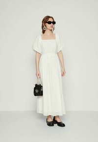Naine kannab valget pitsilised puhv-sleeve maxi kleiti, musti prille, musti kingi, suuri kõrvarõngaid ja hoiab käes musta käekotti lillede detailidega.