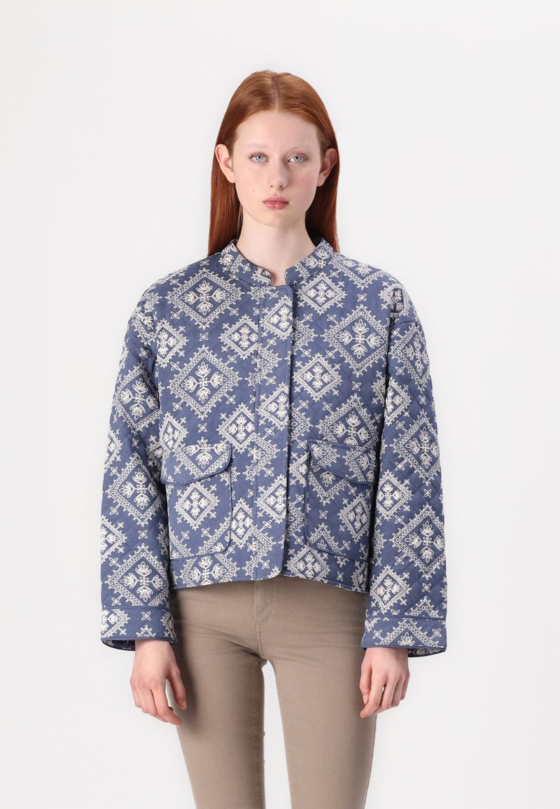 Plavi jakna s quilted uzorkom i bijelim složenim motivima, okrugli izrez i dva prednja džepa. Ima duge, nešto prevelike rukave.