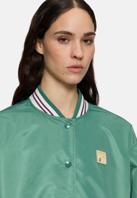 Groene satijnen bomberjack met een gestreepte kraag in wit en paars, voorzien van een gouden geborduurd logo aan de linkerzijde.