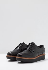 Les chaussures brogues en cuir noir présentent une finition brillante, un bout arrondi, un design à bout ailé et une semelle superposée avec un accent en bois.